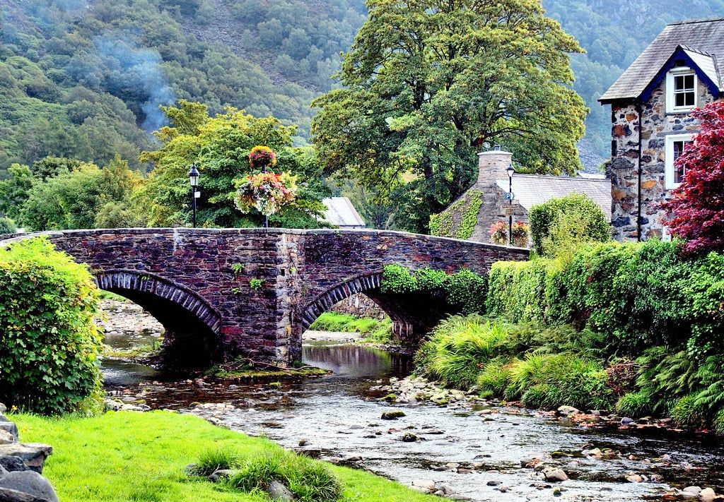 Een historische stenen brug over een rivier, geflankeerd door stenen huisjes en weelderige bomen, in het dorp Beddgelert in Snowdonia.