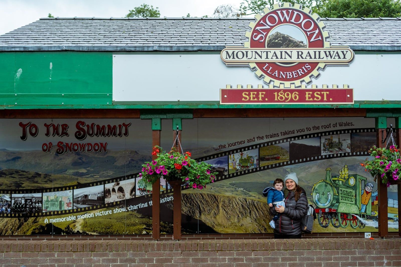 Een vrouw met een jong kind staat voor het stationsbord en muurschildering van de Snowdon Mountain Railway in Llanberis, Snowdonia.