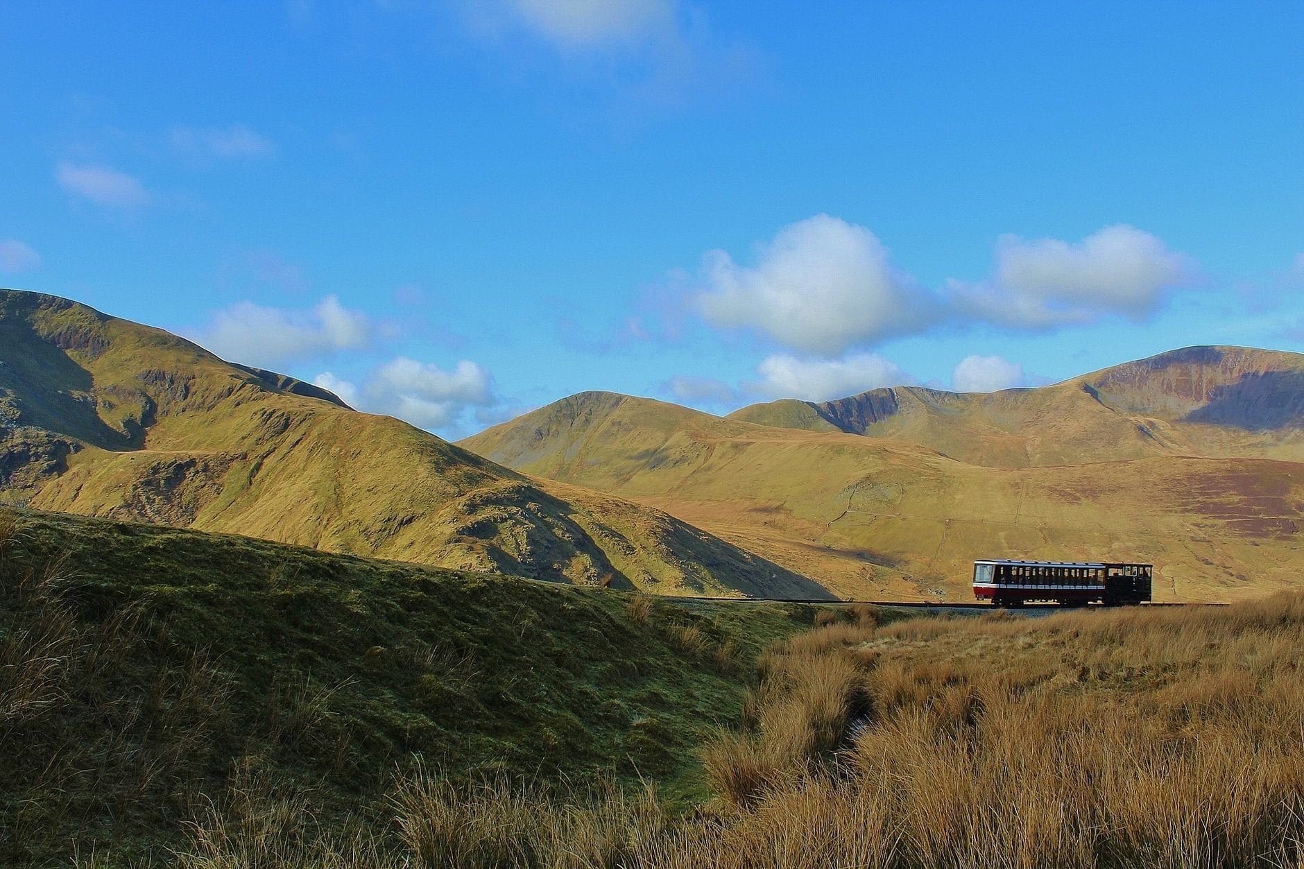 Zug der Snowdon Mountain Railway fährt über die sanften Berge von Snowdonia unter blauem Himmel.