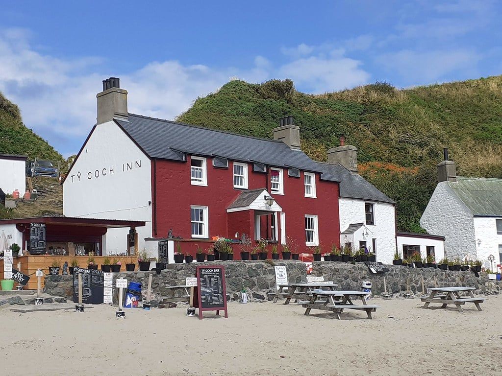 Das Tŷ Coch Inn mit roter Fassade mit Blick auf den Sandstrand mit hölzernen Picknicktischen in Porthdinllaen.