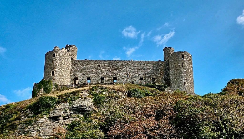 Mittelalterliche Steinmauern und runde Türme von Harlech Castle ragen über einem felsigen, bewachsenen Hügel in Snowdonia unter klarem blauem Himmel auf.