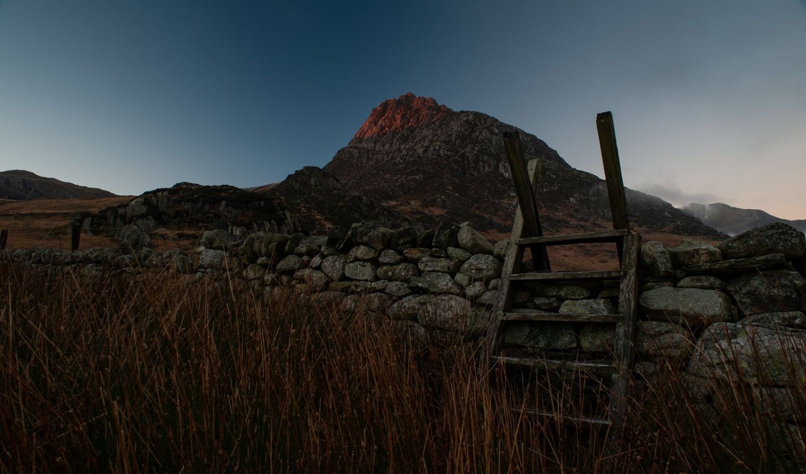Eine hölzerne Leiter über eine Trockenmauer vor dem Gipfel des Tryfan in der Abenddämmerung in Snowdonia.