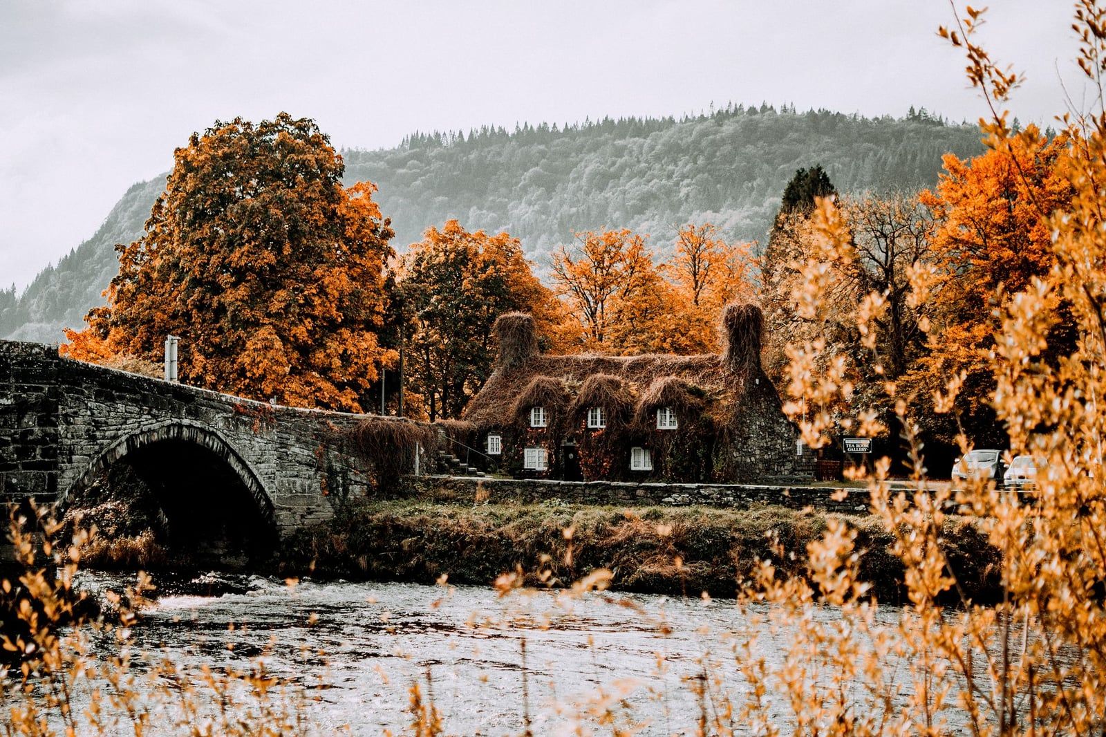 Eine gewölbte Steinbrücke und ein Cottage, gerahmt von leuchtend orangem Herbstlaub am Fluss nahe Betws-y-Coed in Snowdonia.