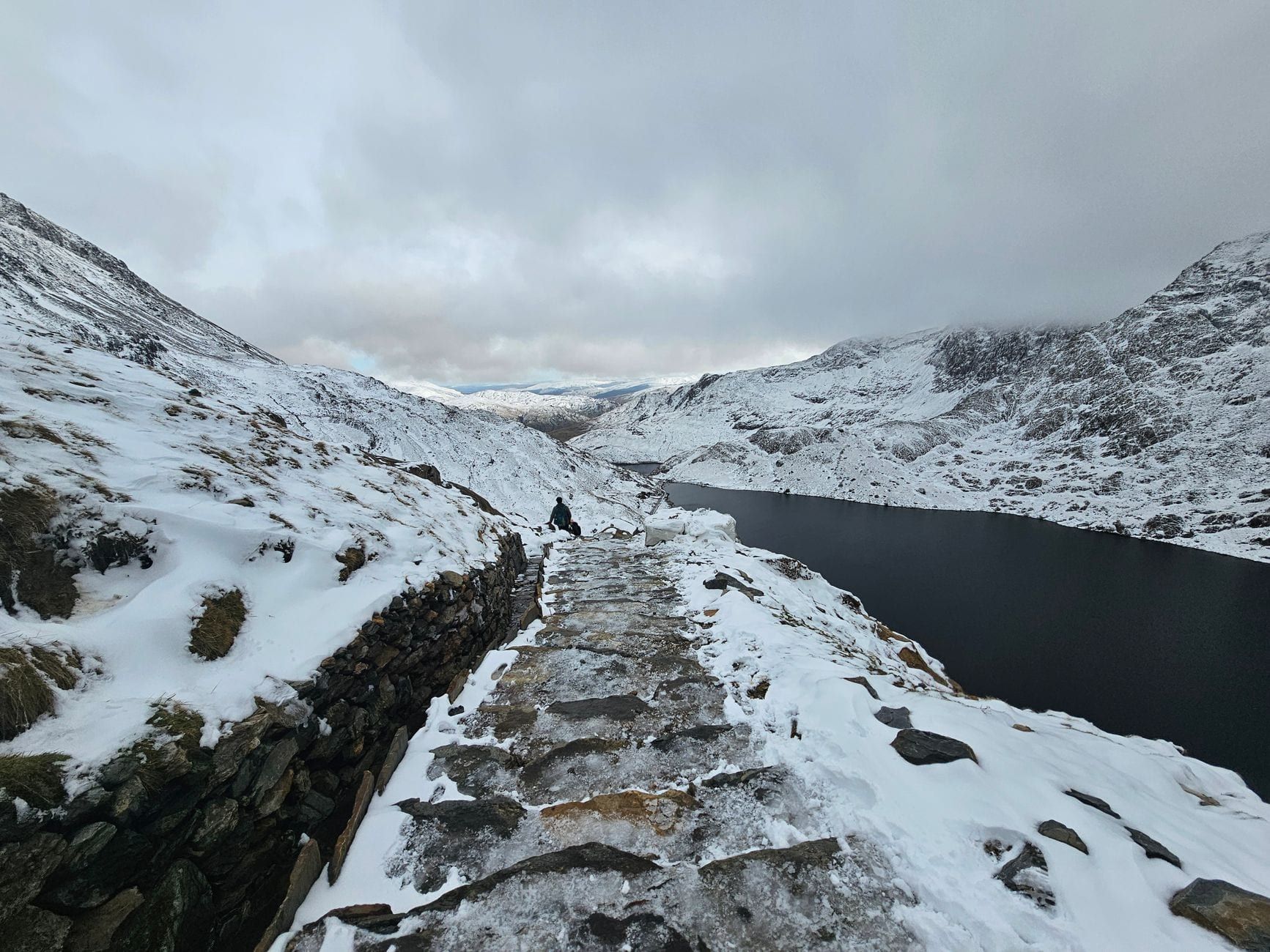 Un sentier de randonnée en pierre recouvert de neige longe une crête montagneuse surplombant un lac dans le Snowdonia.