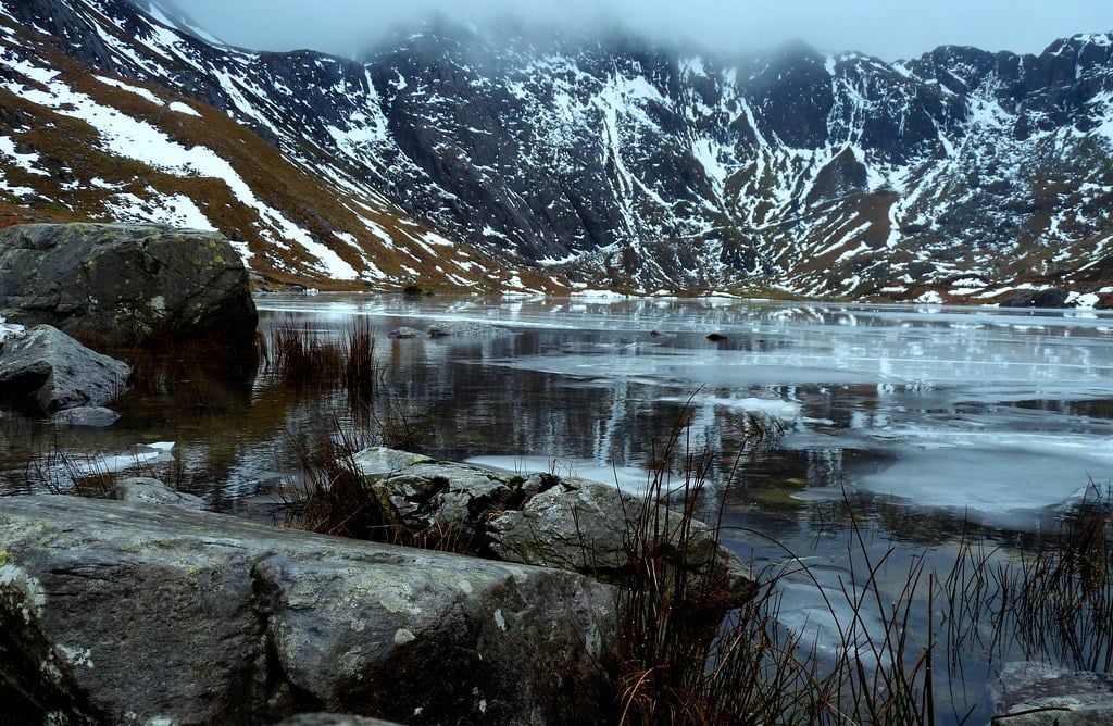 Snow dusted peaks rise above the icy surface of Llyn Idwal in Snowdonia during winter.