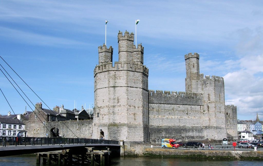 The medieval polygonal towers and curtain walls of Caernarfon Castle stand beside the waterfront under a blue sky near Snowdonia.