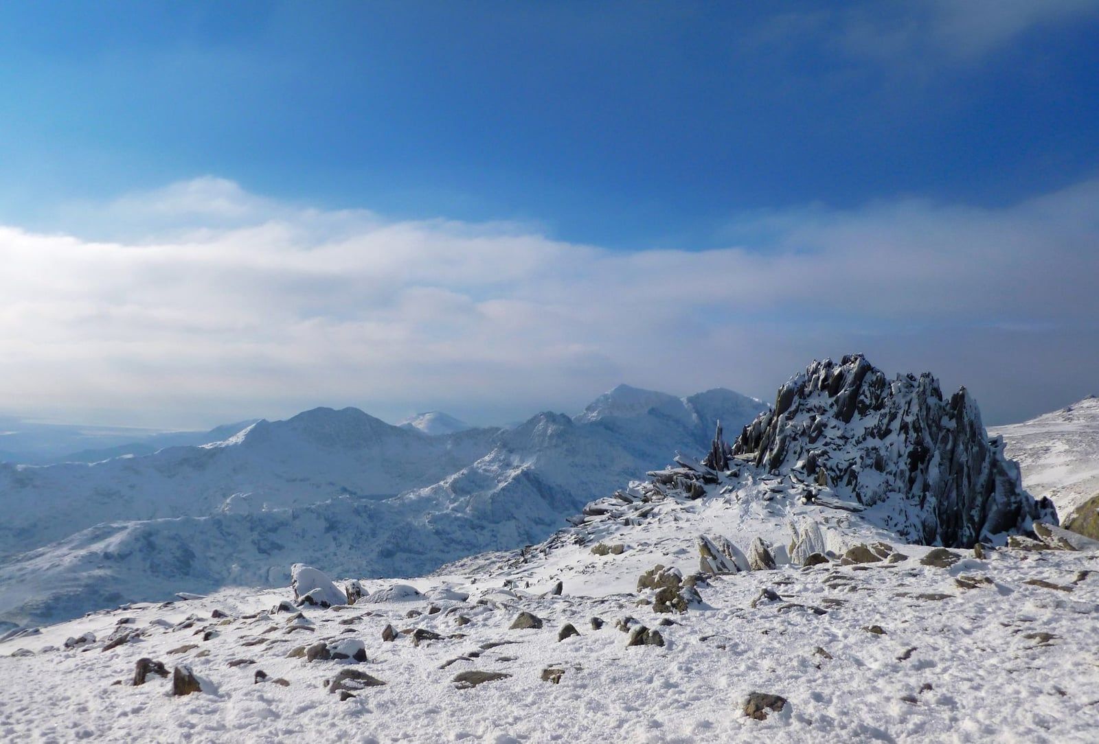 Snow-covered peaks and rugged rocky summits stretch across Snowdonia under a bright winter sky.