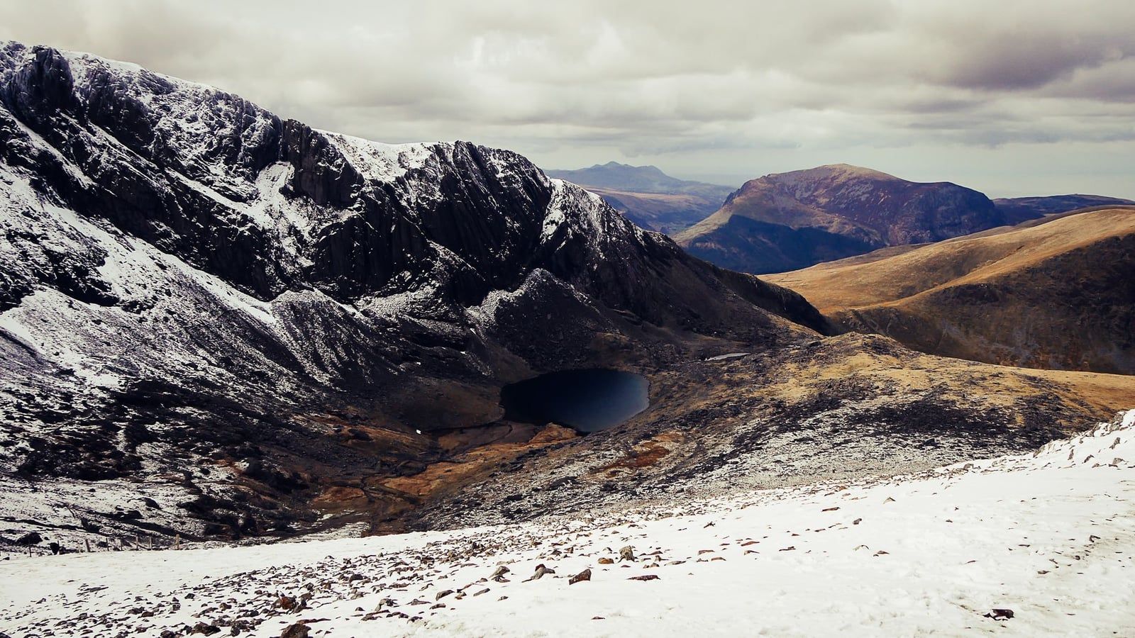 Snow-covered slopes overlooking the dark waters of Llyn Du'r Arddu and rugged mountain ridges in Snowdonia in winter.