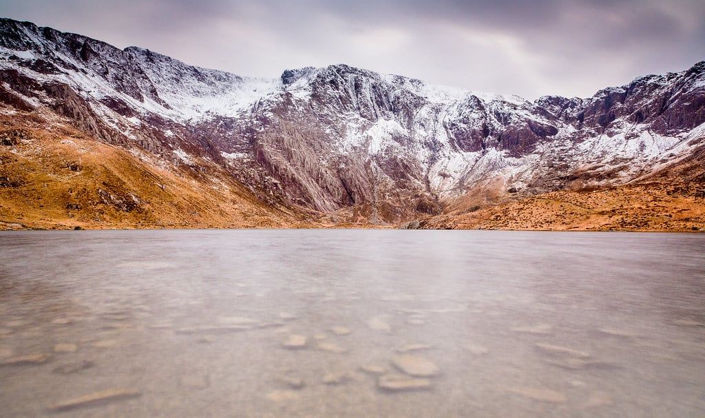 Snow dusts the dramatic cliffs and crags of Cwm Idwal rising above the clear waters of Llyn Idwal in Snowdonia.